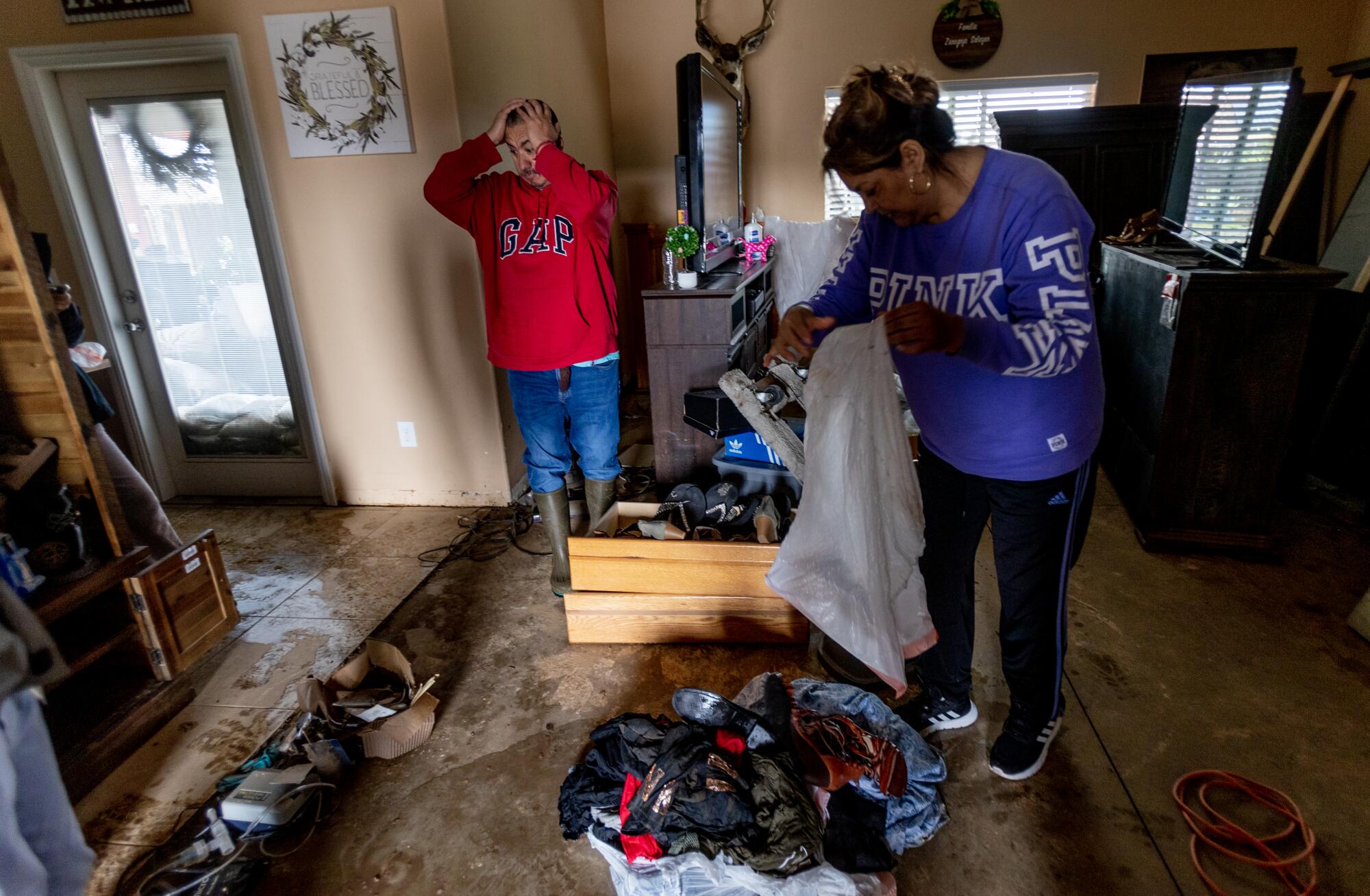 A man grabs his head in frustration while a woman holds a trash bag. Both stand inside a home with muddied, wet floor.