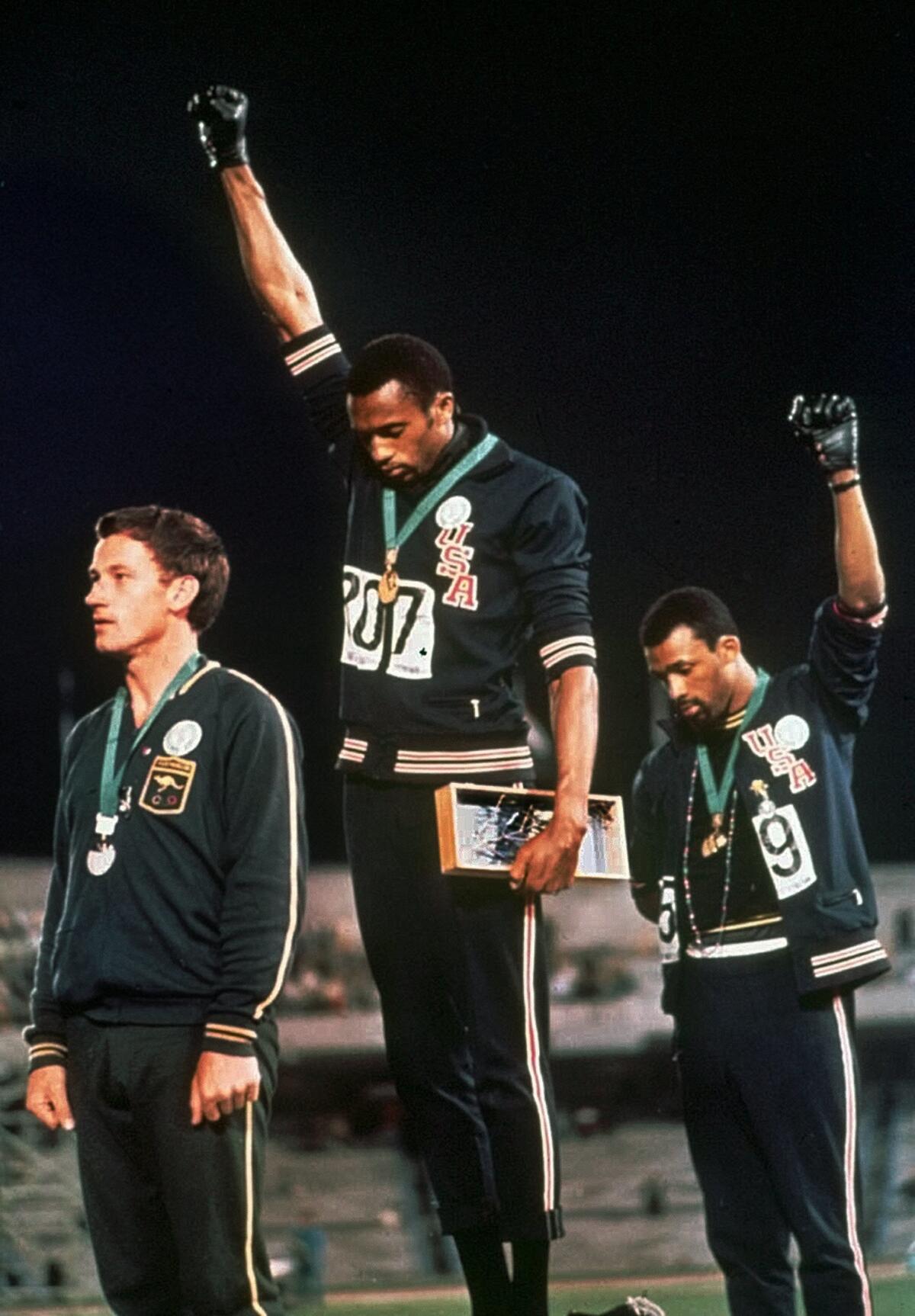 Tommie Smith, center, and John Carlos stare downward on the podium while extending gloved hands skyward.