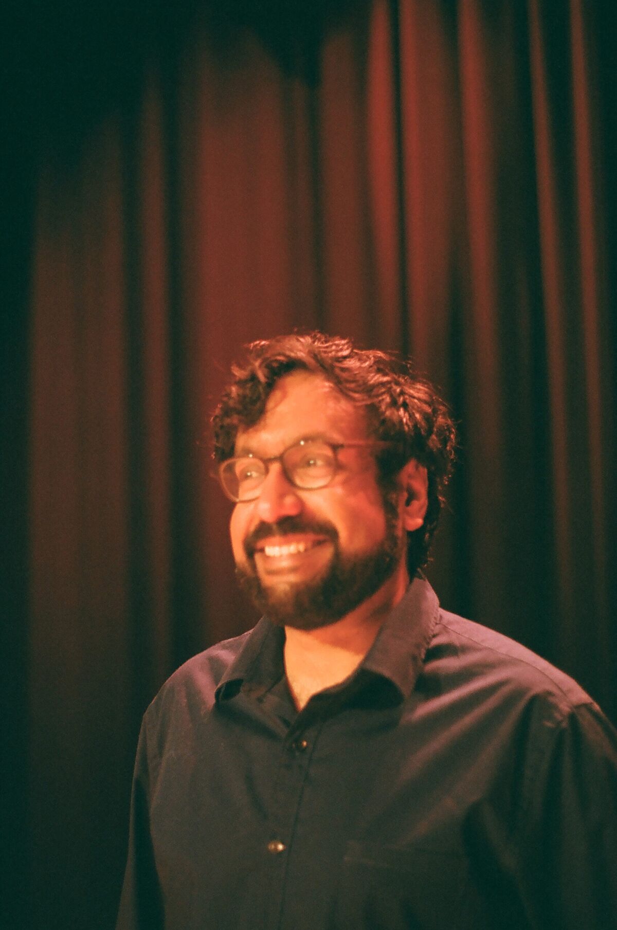 Hari Kondabolu in a black shirt and glasses smiling in front of a red curtain