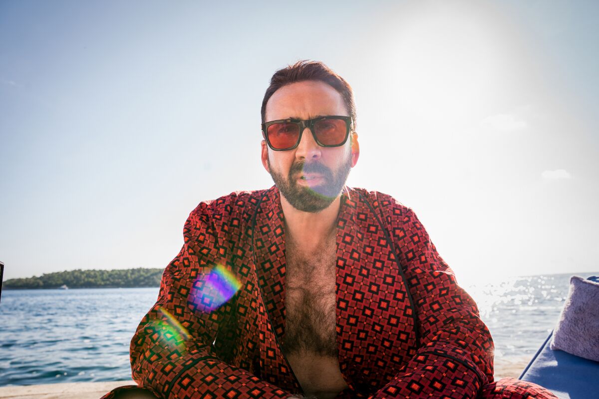 A man in sunglasses and a patterned shirt sits in front of a coastline.