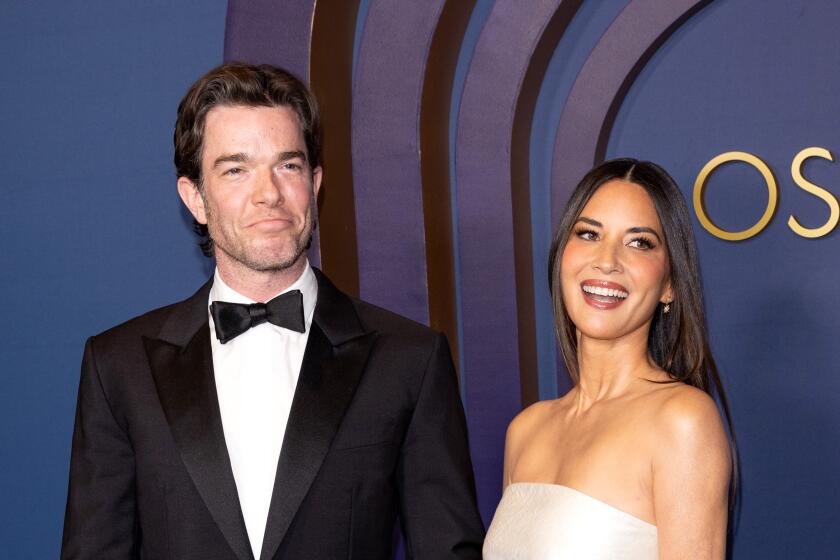 John Mulaney in a tuxedo and Olivia Munn in a strapless white gown smiling as they arrive together at the Oscars