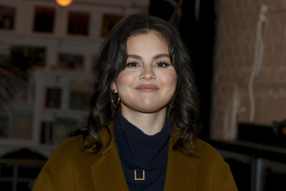 Selena Gomez smiles while wearing a brown coat and a black mock turtleneck