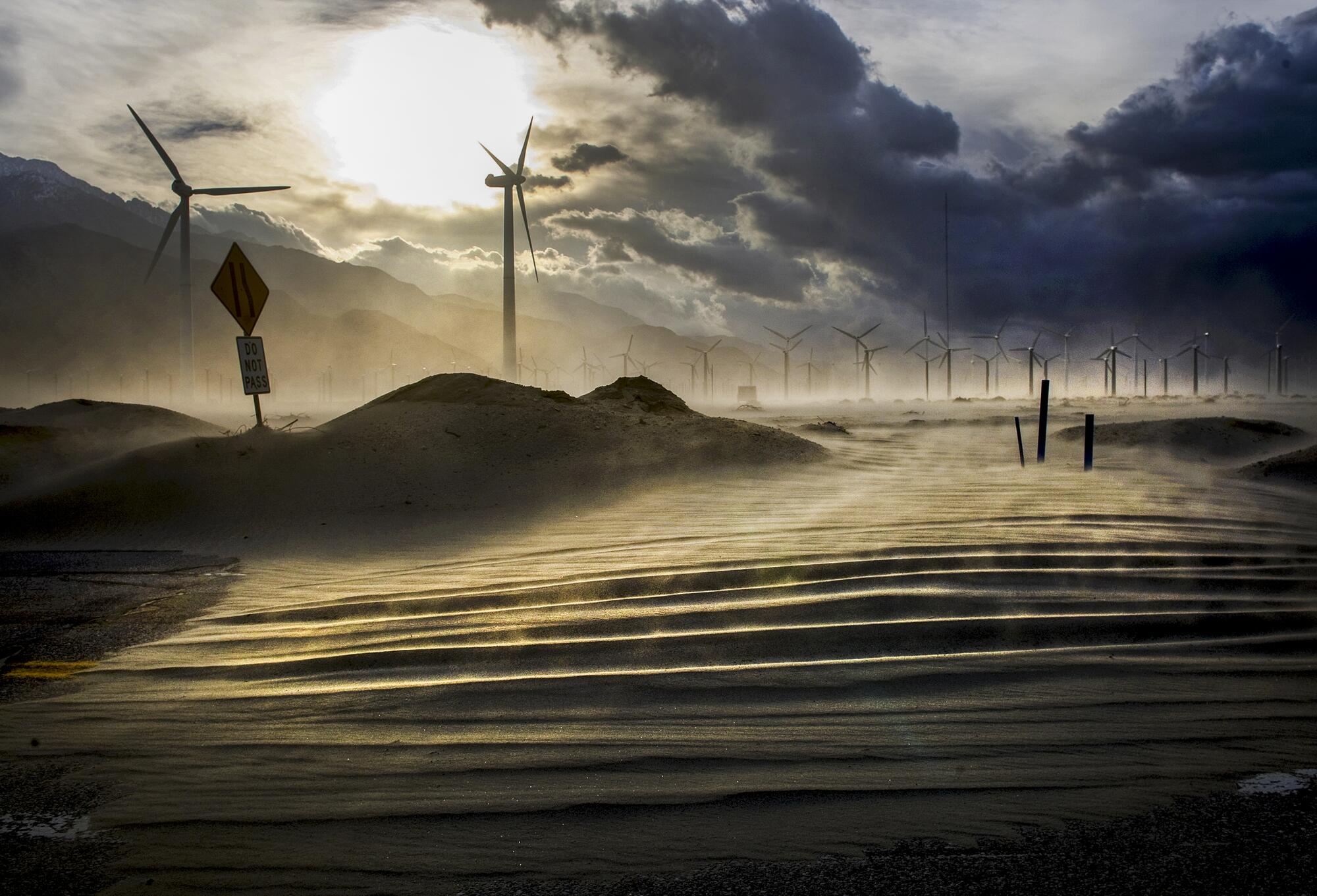 Windmills loom behind a road being covered with drifting sand