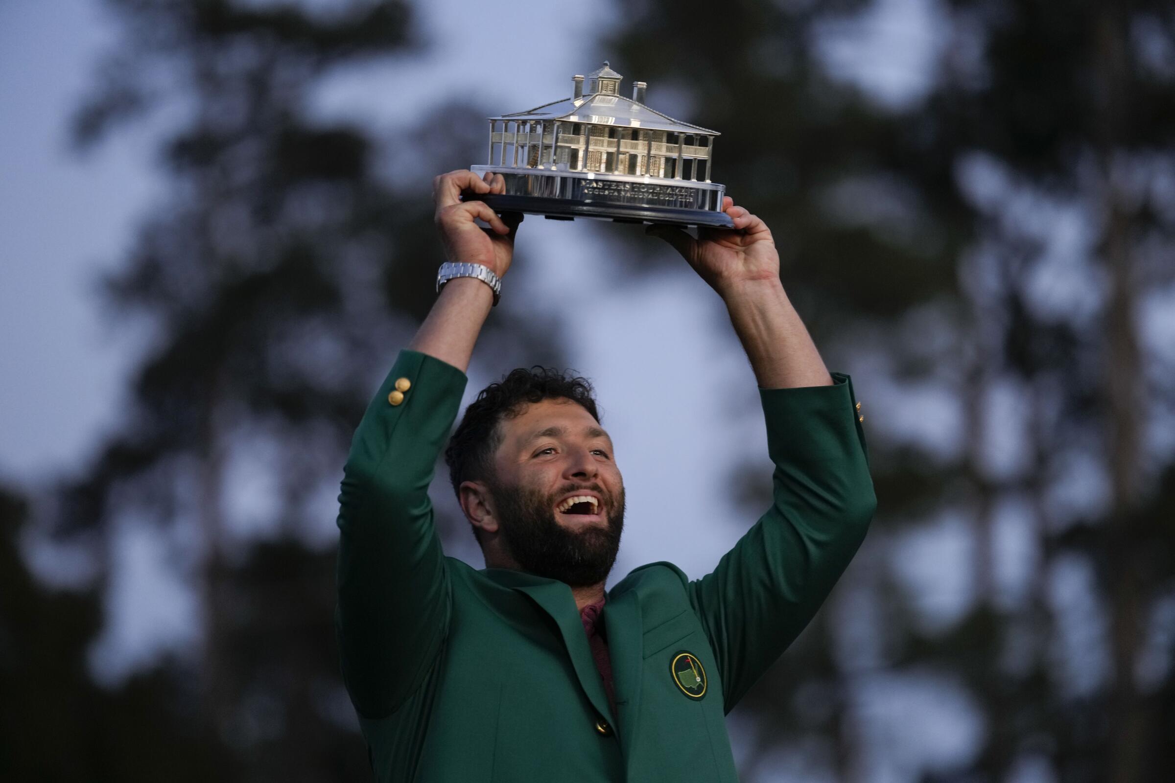 Jon Rahm wears a green jacket and holds the Masters trophy above his head.