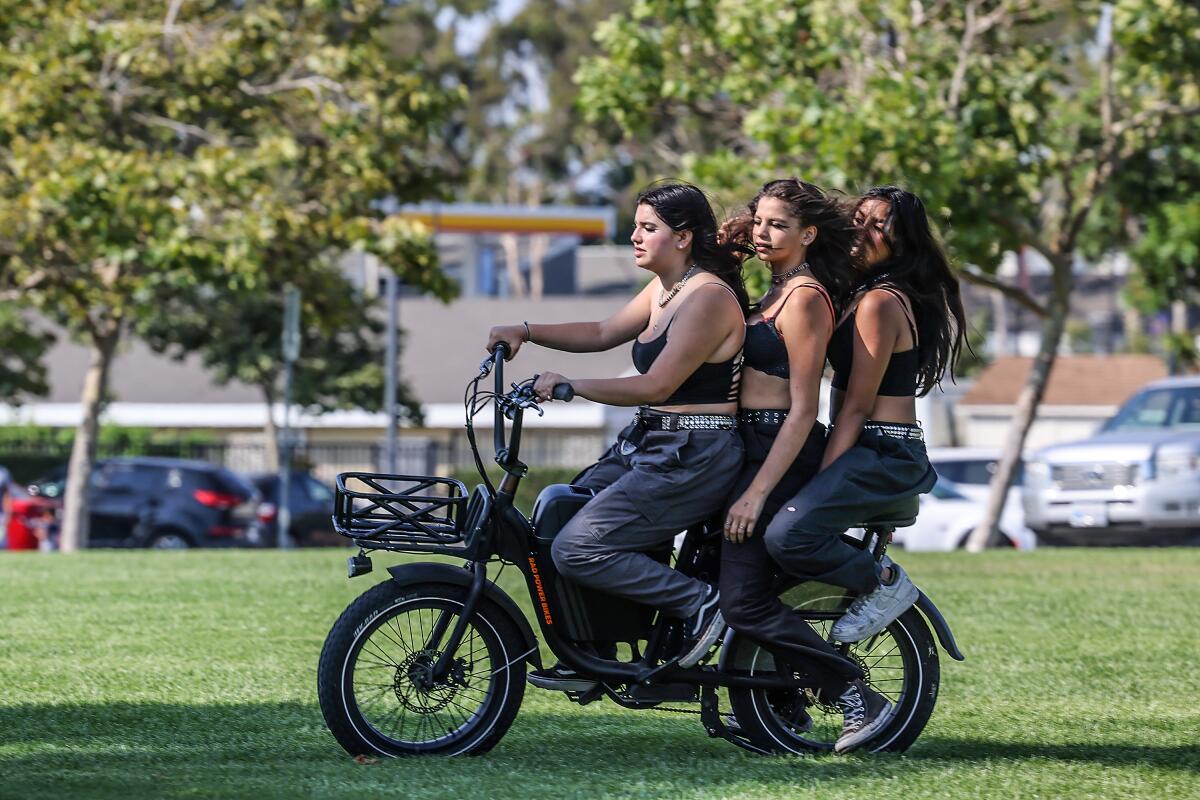 Three women ride one e-bike on the grass at Mike Ward Community Park in Irvine in July 2021.