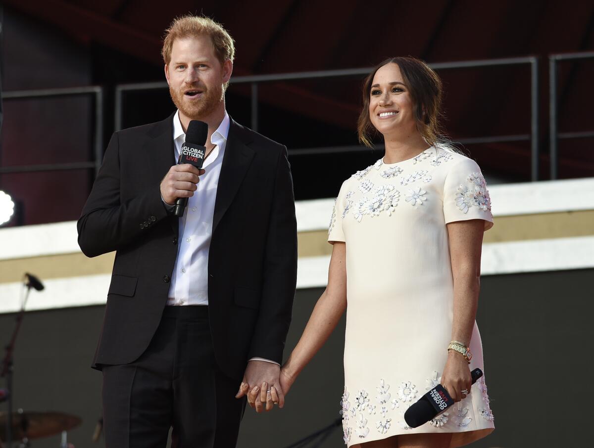 Prince Harry in a black blazer and pants standing next to and holding hands with Meghan Markle in a white dress 