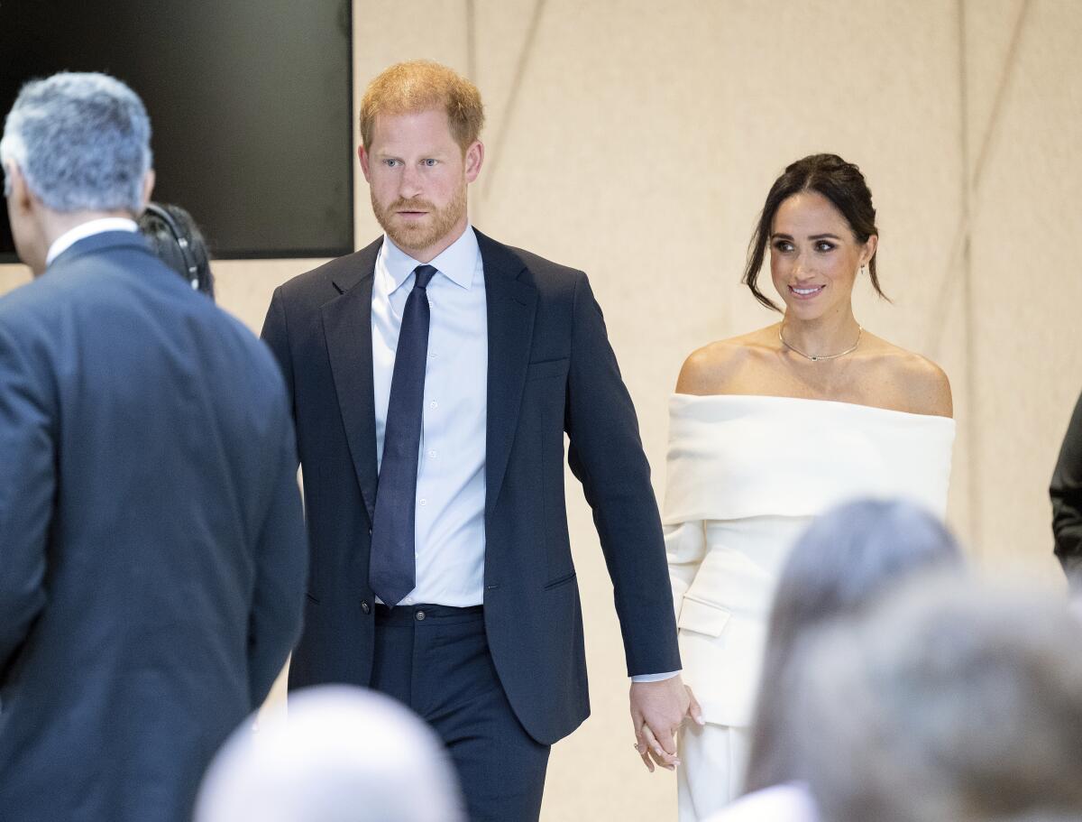 Prince Harry in a suit and Meghan in a white dress hold hands as they walk into a room toward people