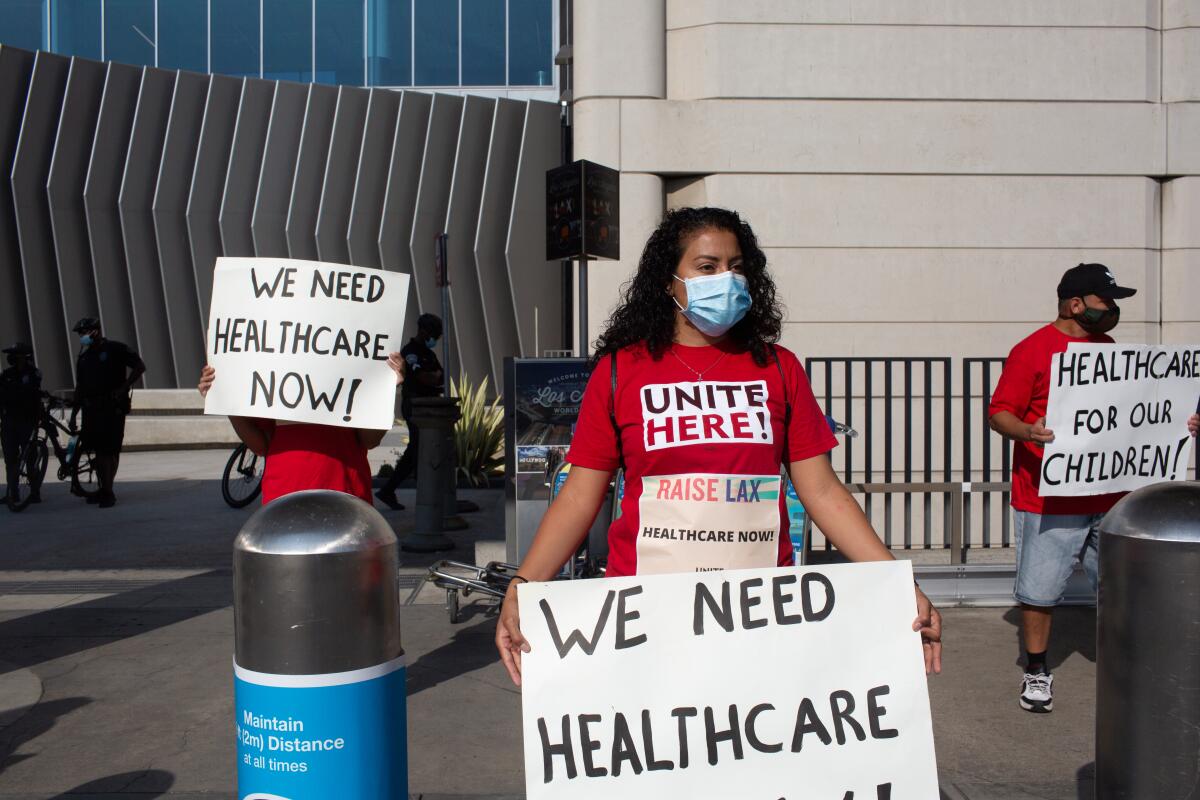 People at Los Angeles International Airport hold signs that say "We need healthcare now!" and "Healthcare for our children!"