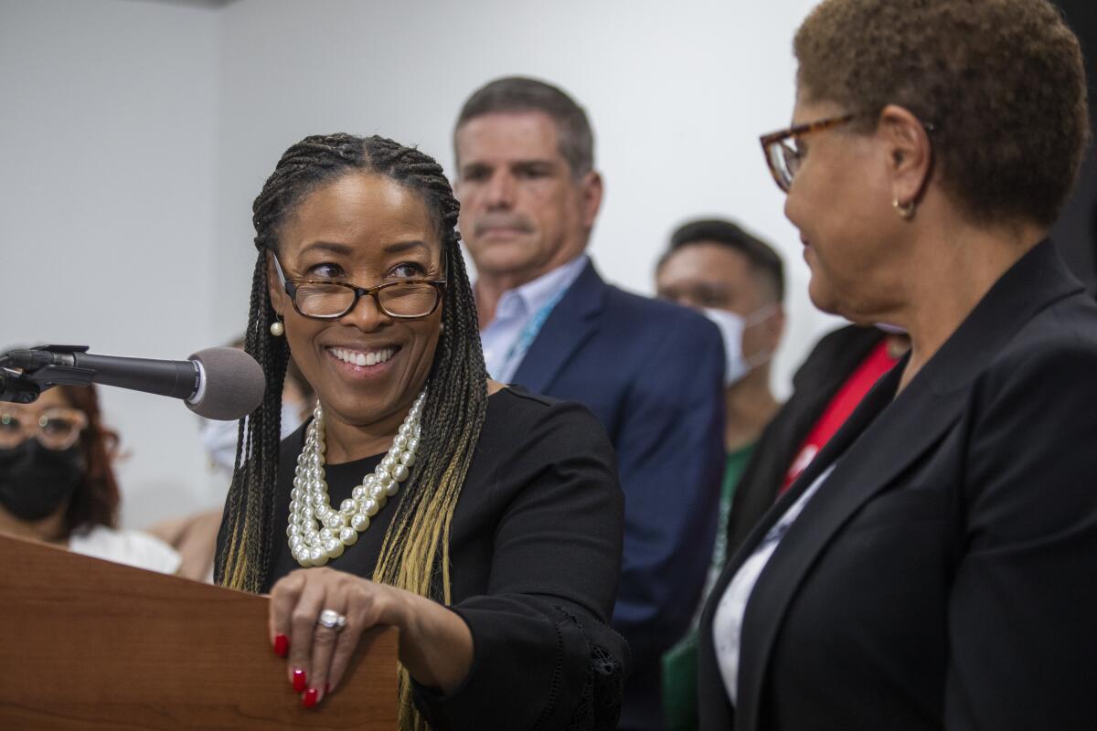 Va Lecia Adams Kellum stands at a microphone and smiles at Mayor Karen Bass