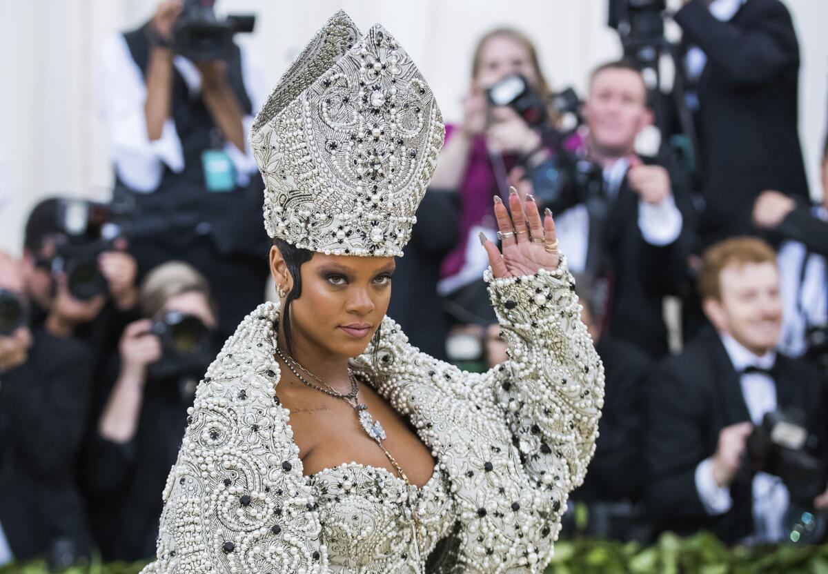 A woman waving while wearing a large bejeweled ecclesiastical-looking hat and matching coat, dress and necklace