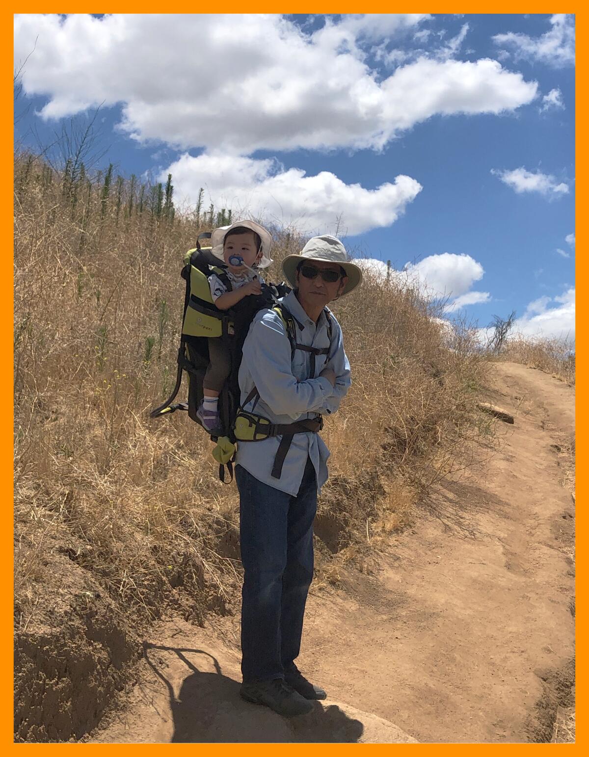 A man in a hat stands with a baby in a carrier on his back on a dirt path under a blue sky with puffy white clouds.