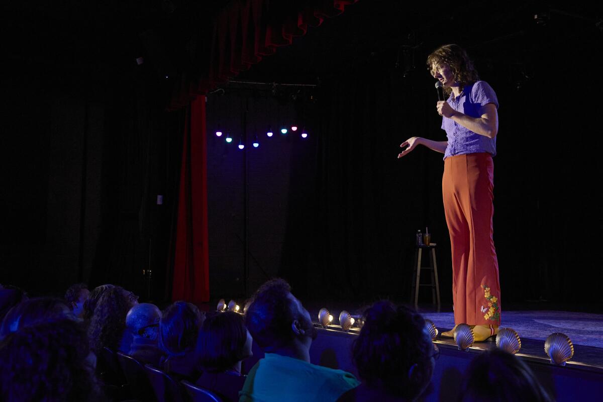 A comedian stands onstage speaking into a microphone in a dark theater 