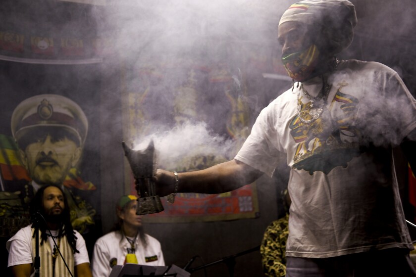 From left, Mosiyah Tafari and Binghi Neal take a break from traditional nyabinghi drumming and chanting on stage as frankincense wafts from a burner held by Ras Jahbo, center, during an event by the Rastafari Coalition marking the 91st anniversary of the coronation of the late Ethiopian Emperor Haile Selassie I in Columbus, Ohio on Tuesday, Nov. 2, 2021. As public opinion and policy continues to shift in the U.S. and across the world towards the use of marijuana, some adherents of Rastafari question their place in the future of the herb that they consider sacred. (AP Photo/Luis Andres Henao)