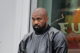 Kanye West poses with a glum look with his hands folded in front of him while wearing a shiny leather coat