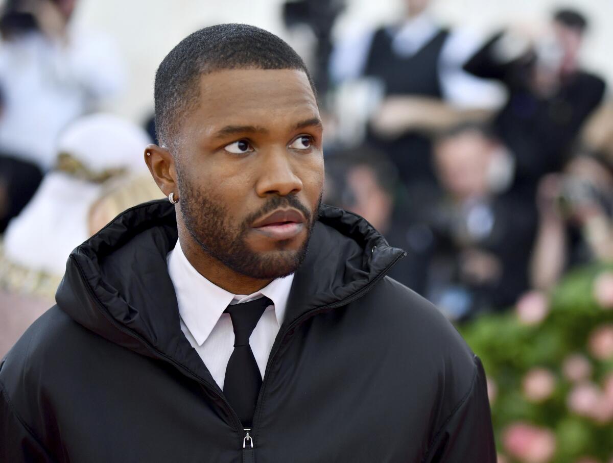 Frank Ocean wears a black coat, white shirt and black tie while staring off into the distance.