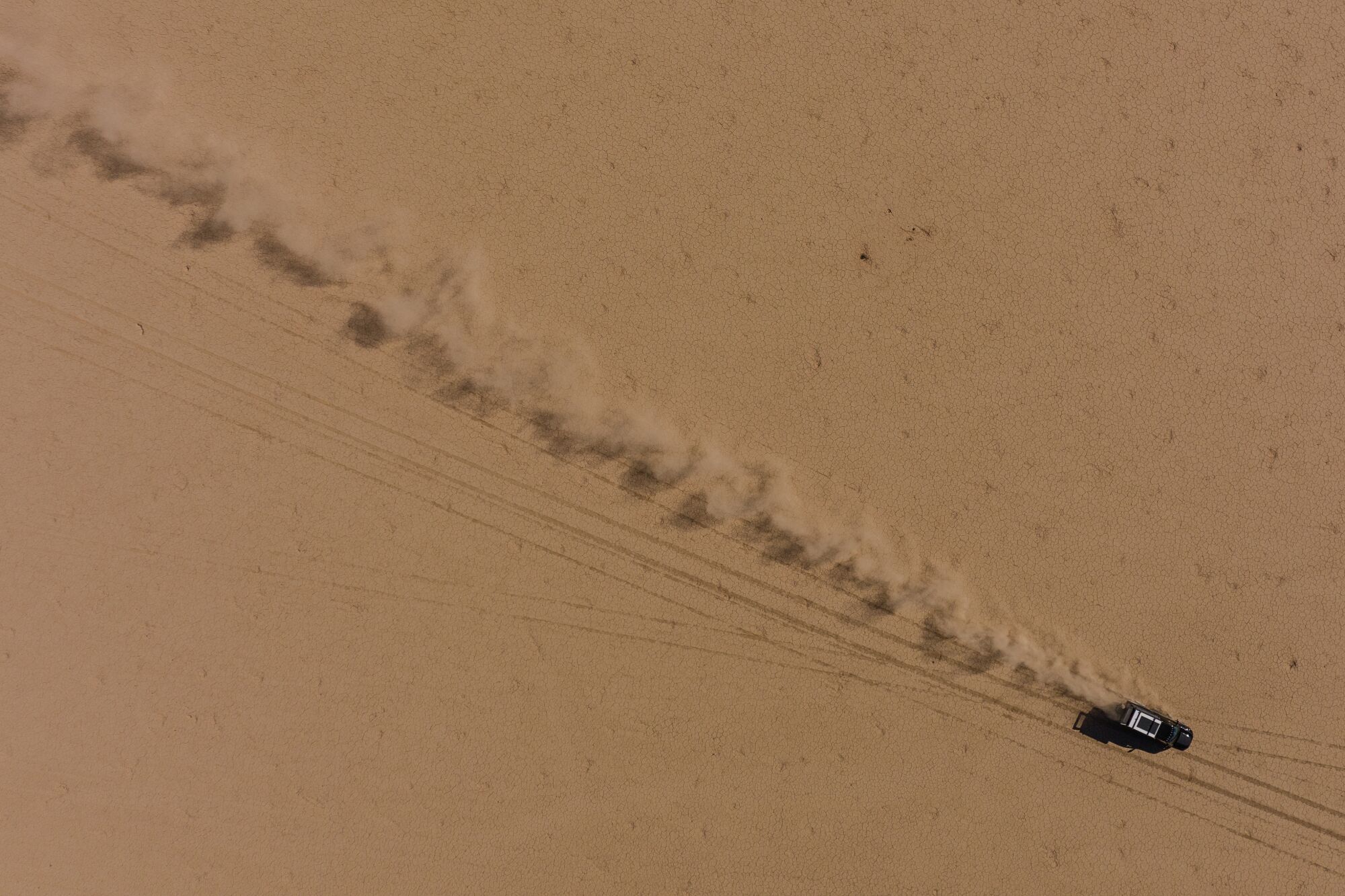 A drone photo of a truck driving across a dry lake bed, kicking up a stream of dust in its wake.