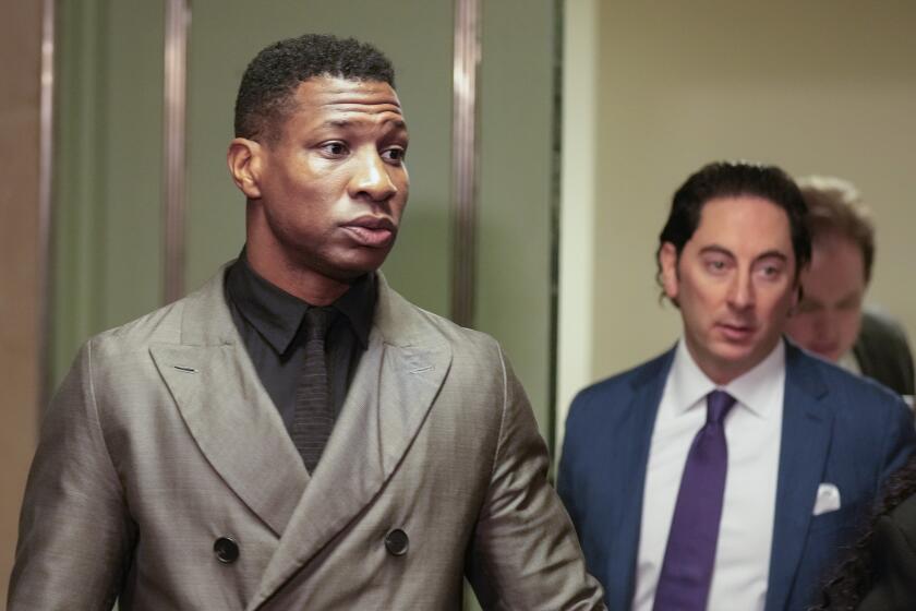 Jonathan Majors standing in front of a door in a grey suit looking to his left in front of a man in a blue suit jacket 