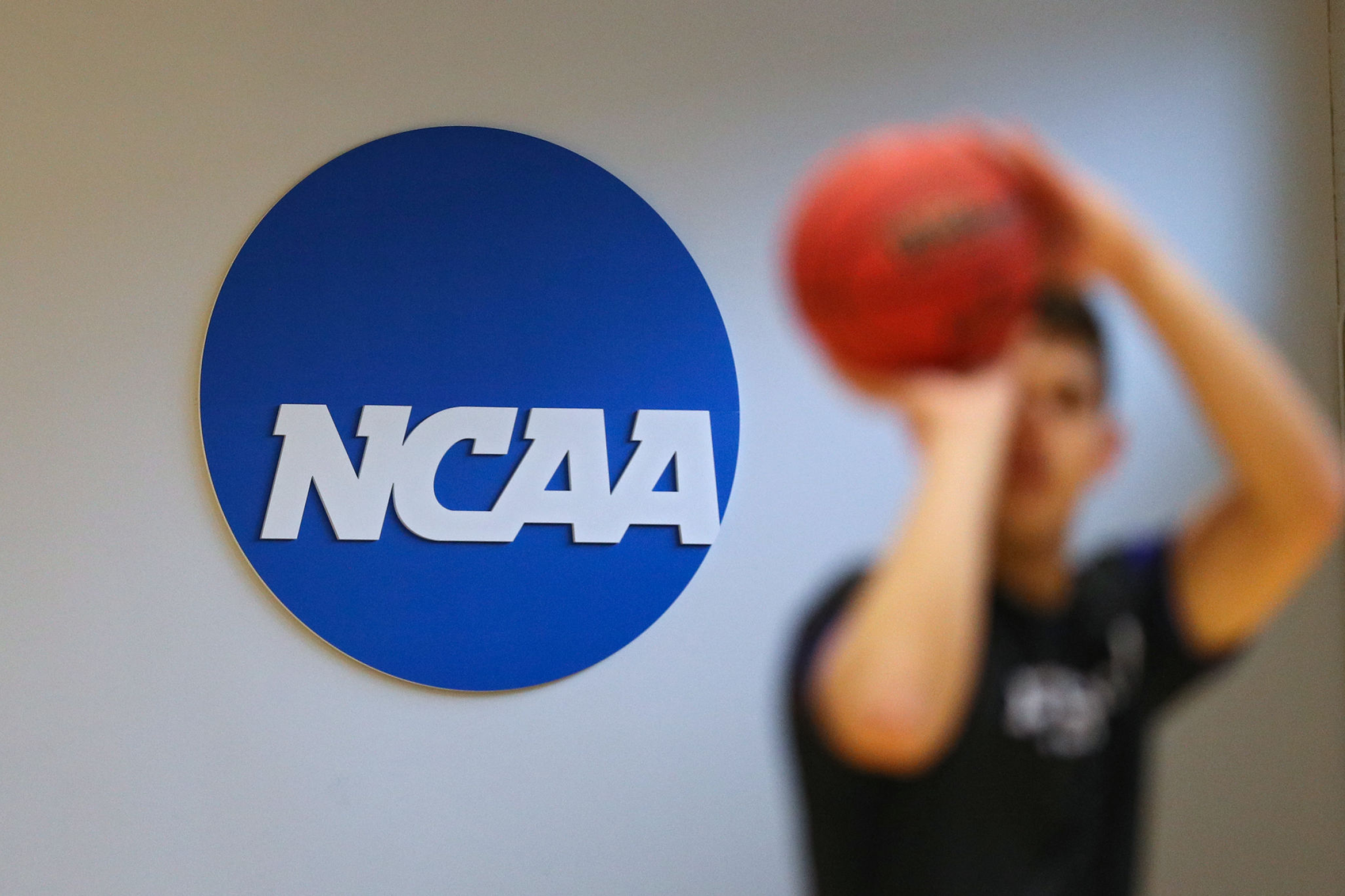 A basketball player shoots in front of the NCAA logo