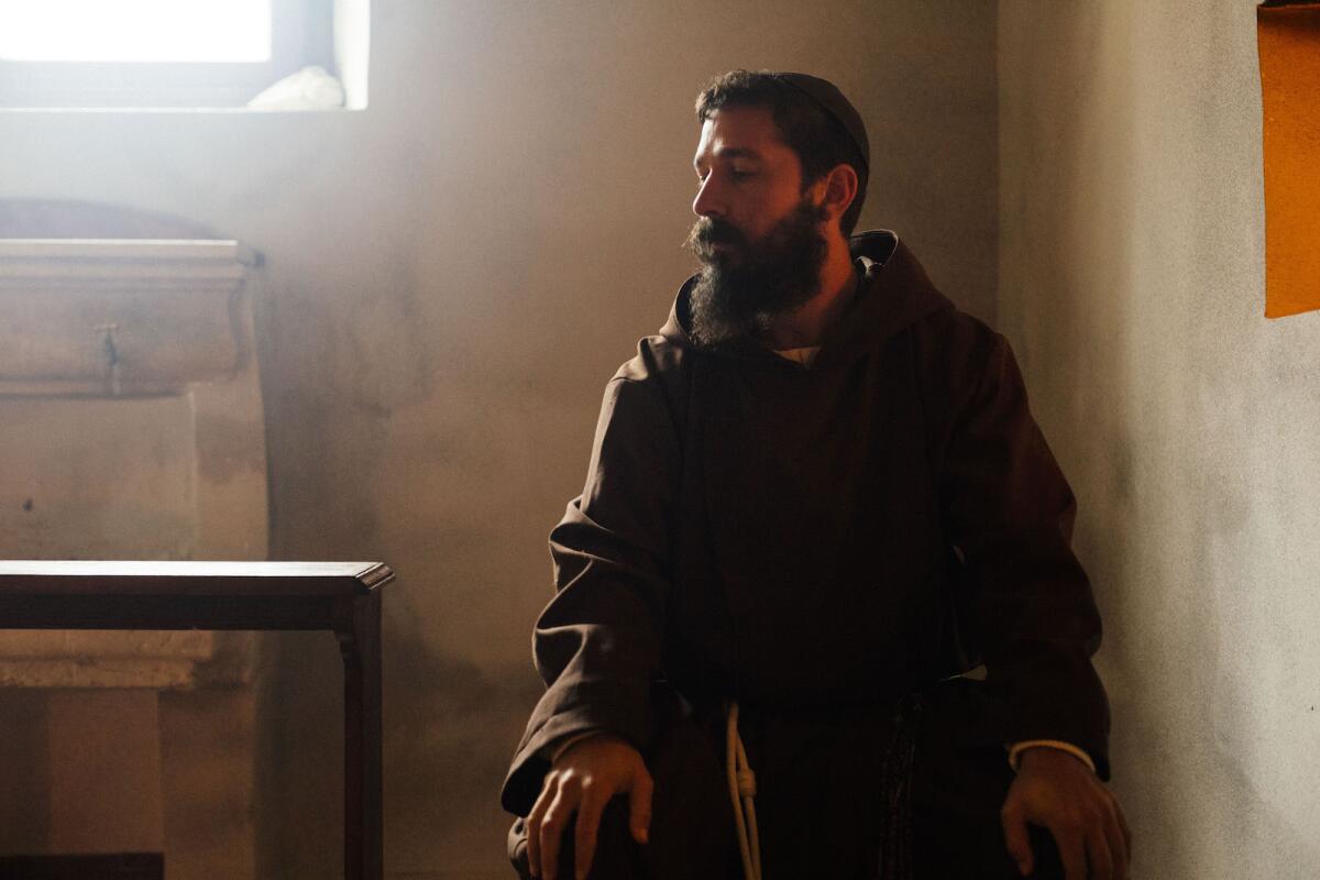 Shia LaBeouf with a thick beard wears a dark brown liturgical vestment as sun shines through a window in a scene from a film