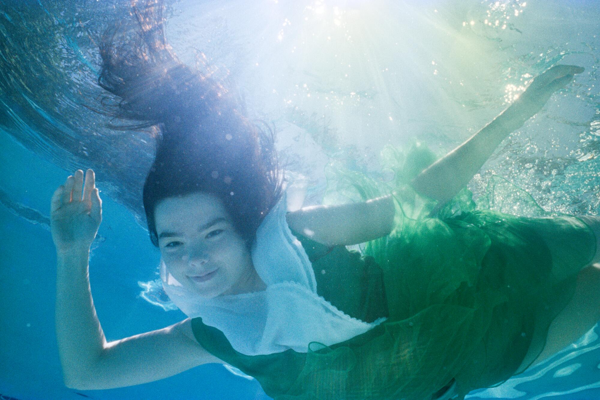 An underwater photo of a smiling woman in a pool with dark hair, wearing a green dress with a white collar