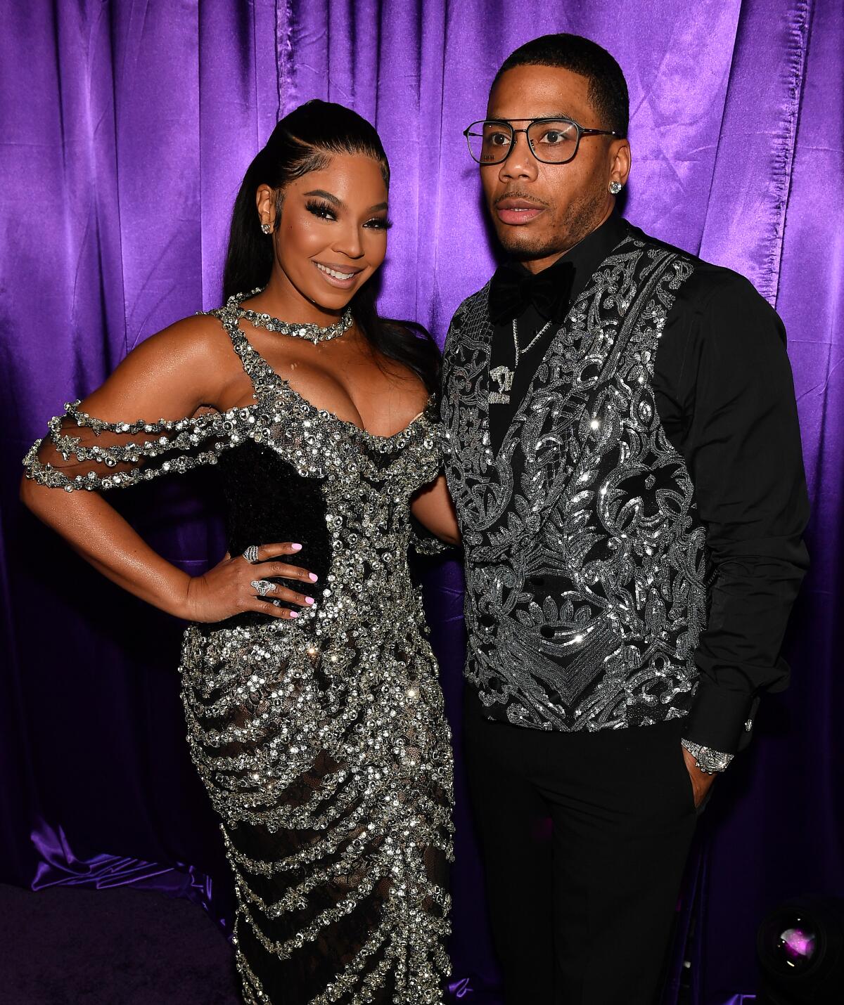 Ashanti smiles in a glittery gown while standing with her arm around rapper Nelly in a glittery vest