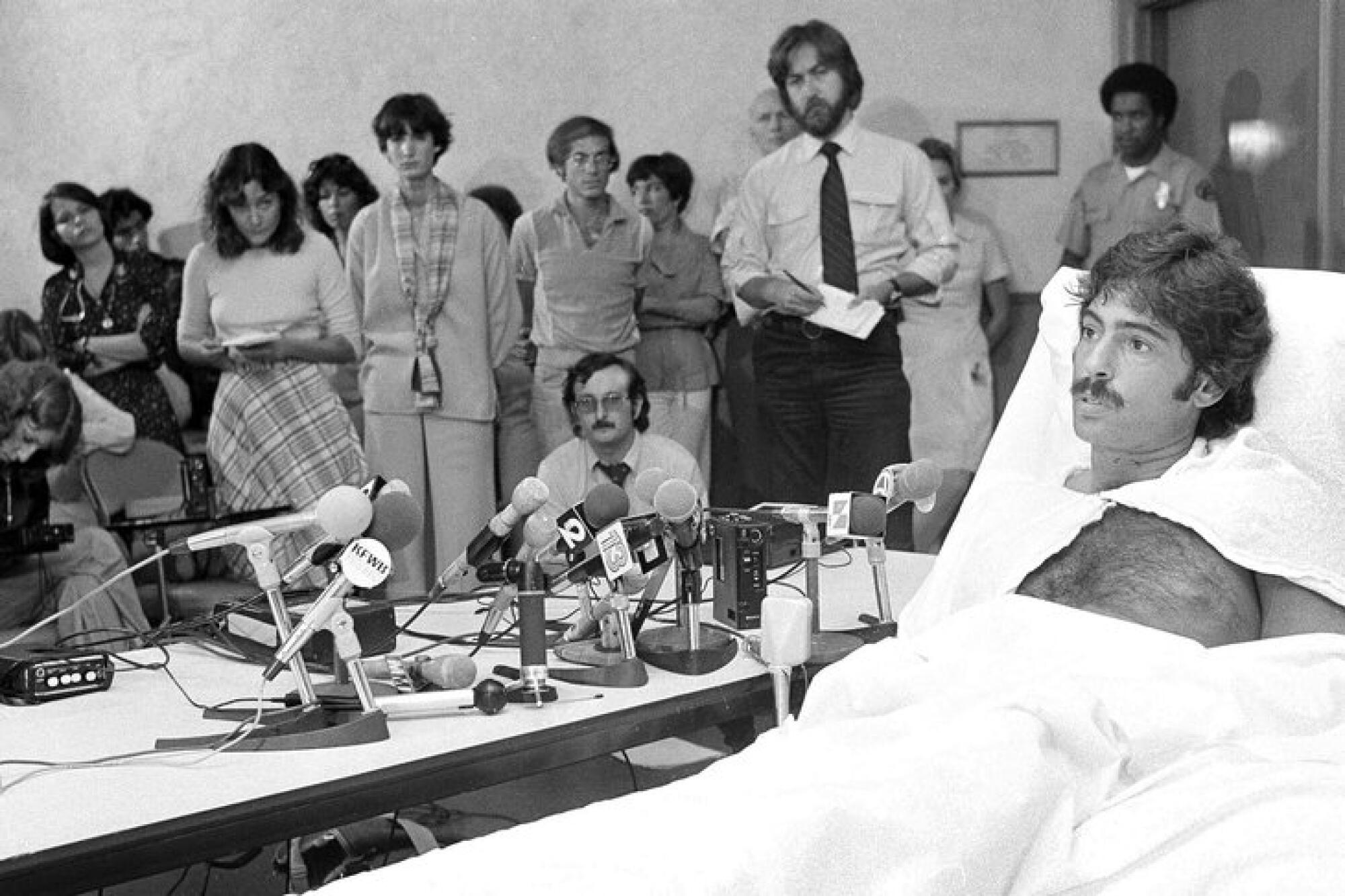 A man in a hospital bed is next to a desk full of microphones, with people standing in the background.