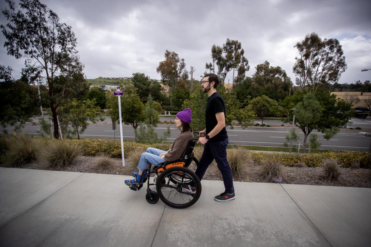 A man pushes a woman in a wheelchair on a sidewalk.