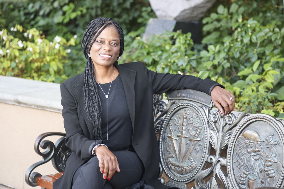 Delicia Turner Sonnenberg wears braids and a black suit, sitting on a bench outside, outside the Globe