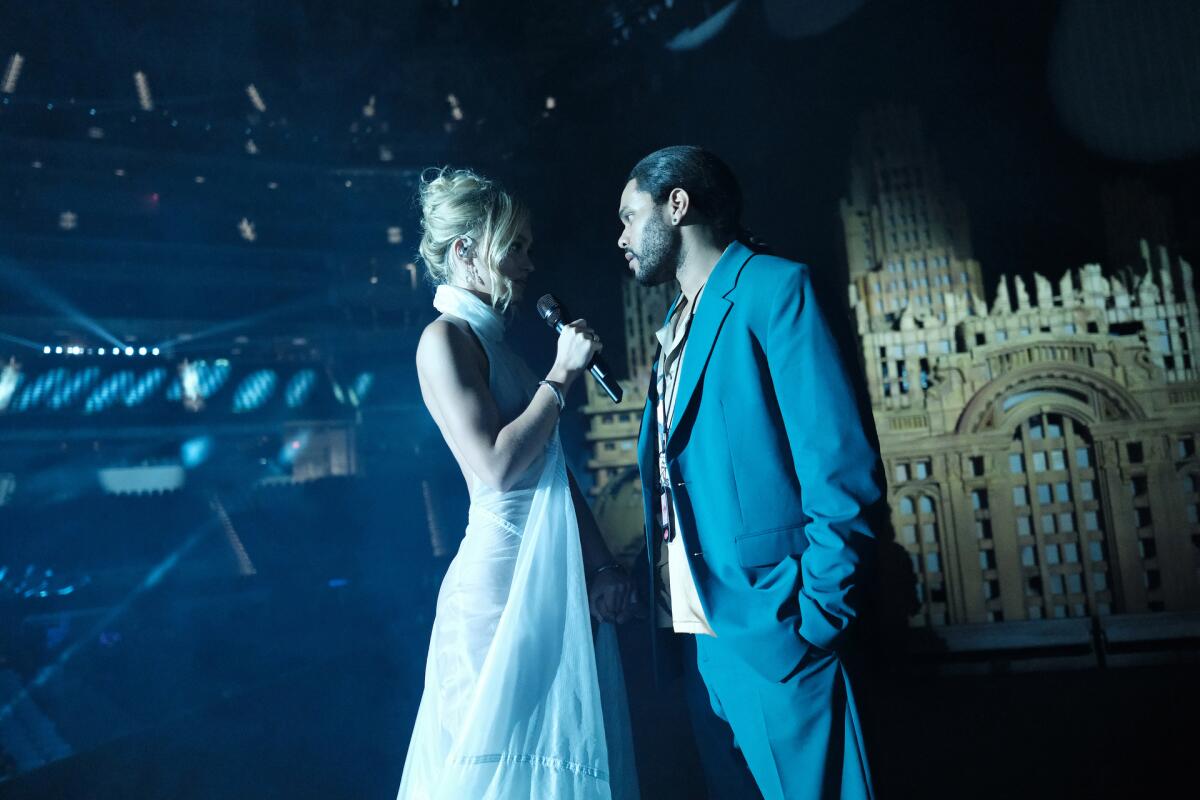 Lily-Rose Depp, wearing a white dress and holding a microphone, faces Abel "The Weeknd" Tesfaye in a suit