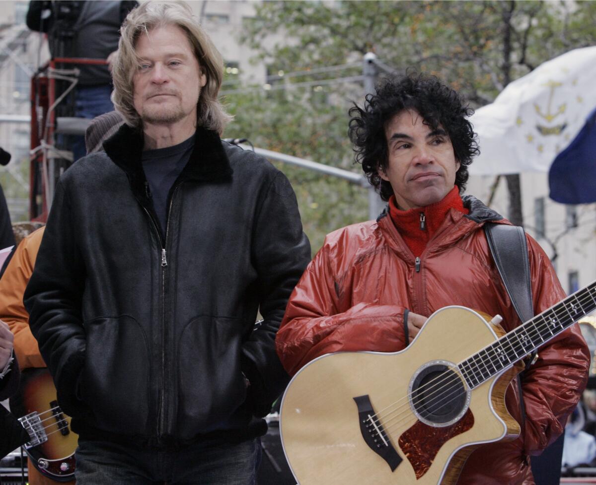 Daryl Hall, left, wears a black jacket and John Oates, right, wears a red jacket and holds a guitar as both stand outdoors