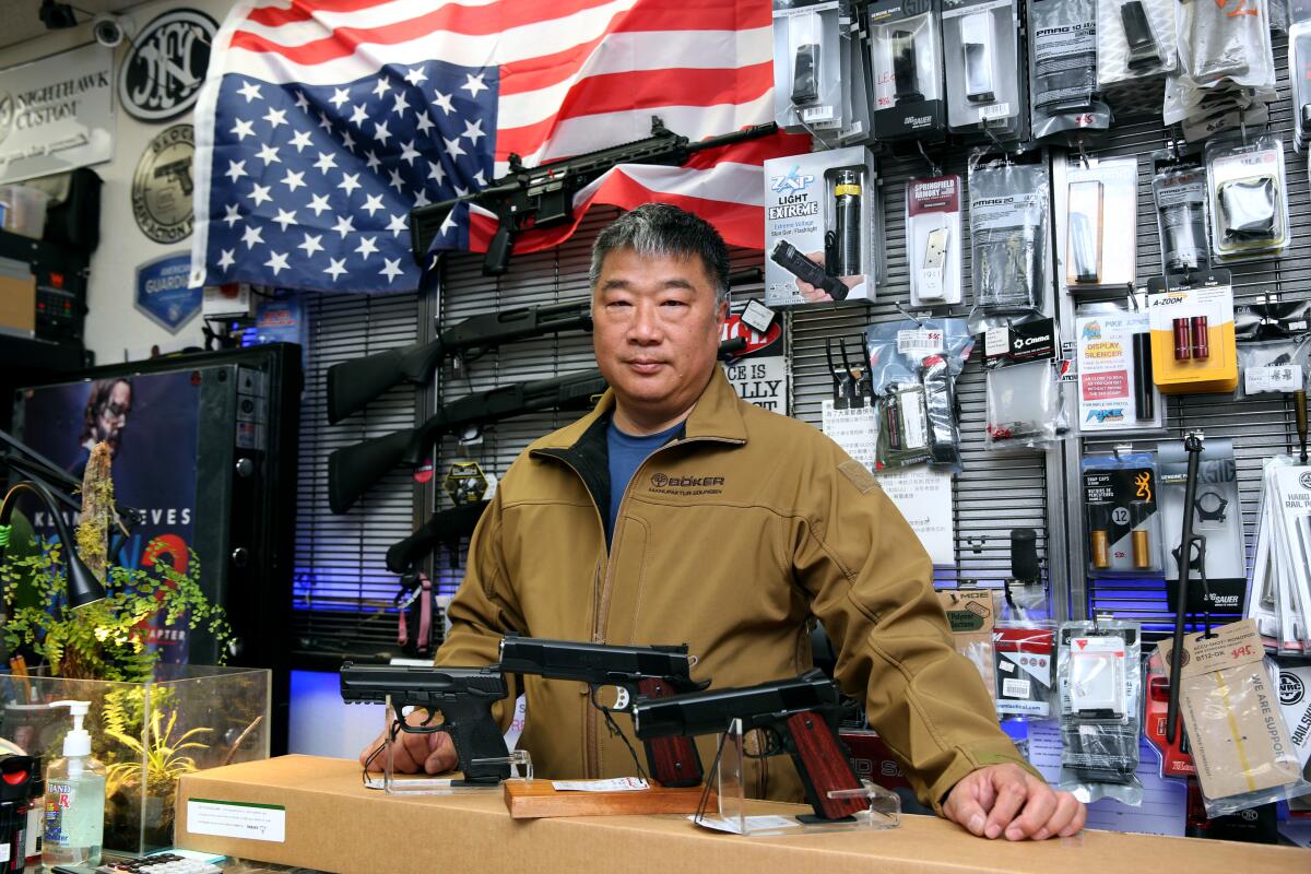 A man standing behind a counter of a gun store with pistols, rifles and packaged accessories on display
