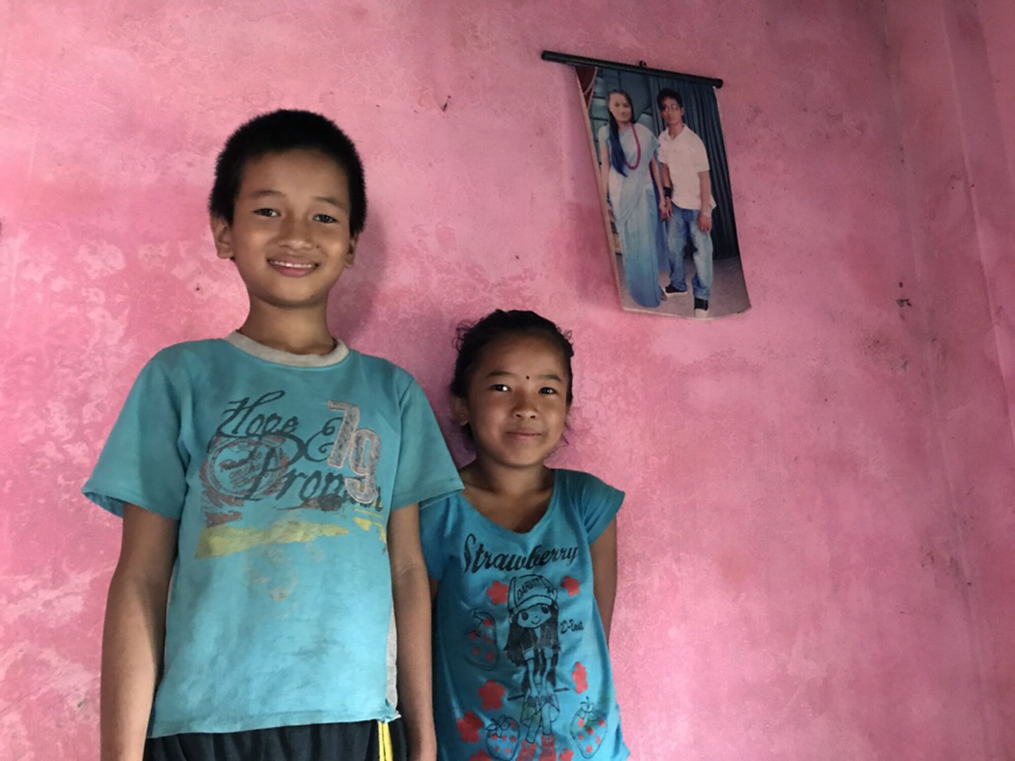 Two siblings stand in front of a pink wall with a photo of their parents on it