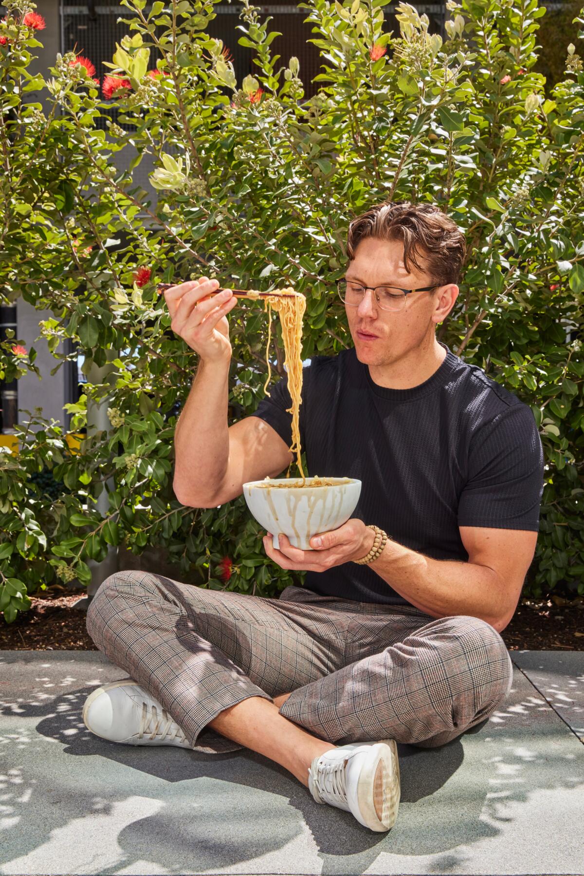 A man sits cross-legged on the ground outdoors, lifting ramen from a bowl with chopsticks