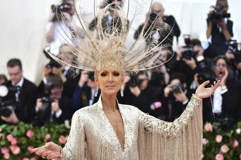 Celine Dion at wearing a gold gown with fringe sleeves, a large matching, feathered headpiece with her arms to the side
