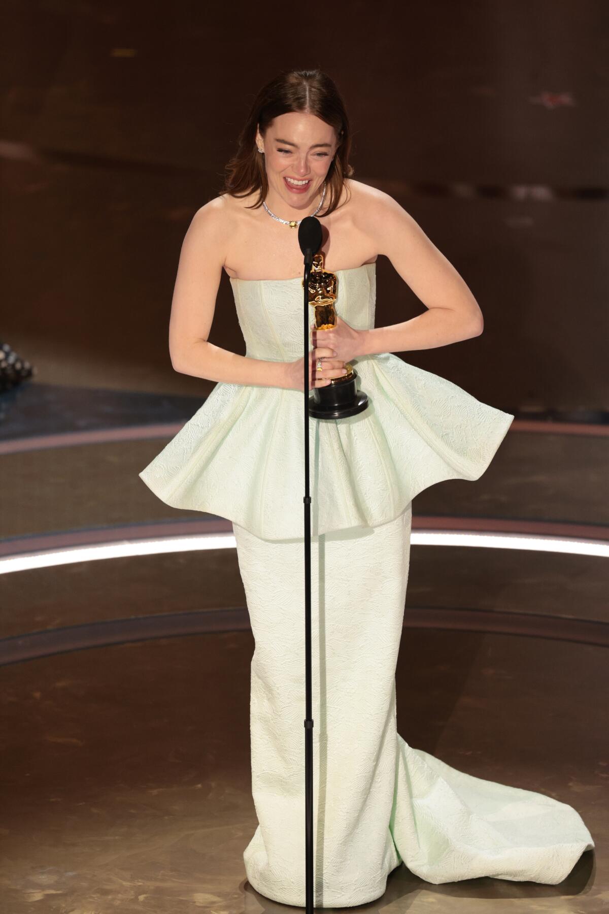 Emma Stone holds an Oscar statue with both hands as she speaks into a mic on stage. 