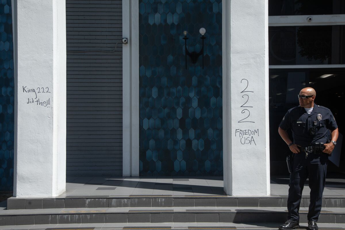 A police officer stands with his hands on his hips outside a mosque with graffiti on its pillars.