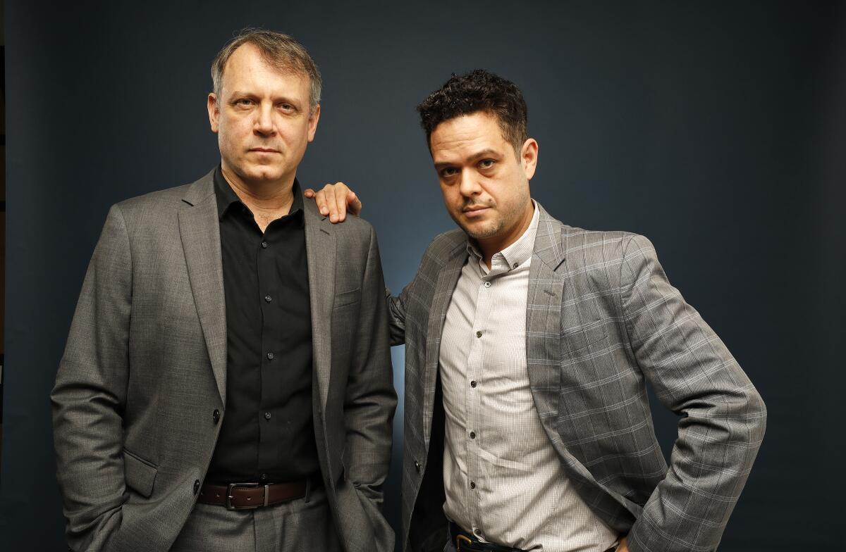 Two men wearing suit jackets pose, one with his hand on the other's shoulder.