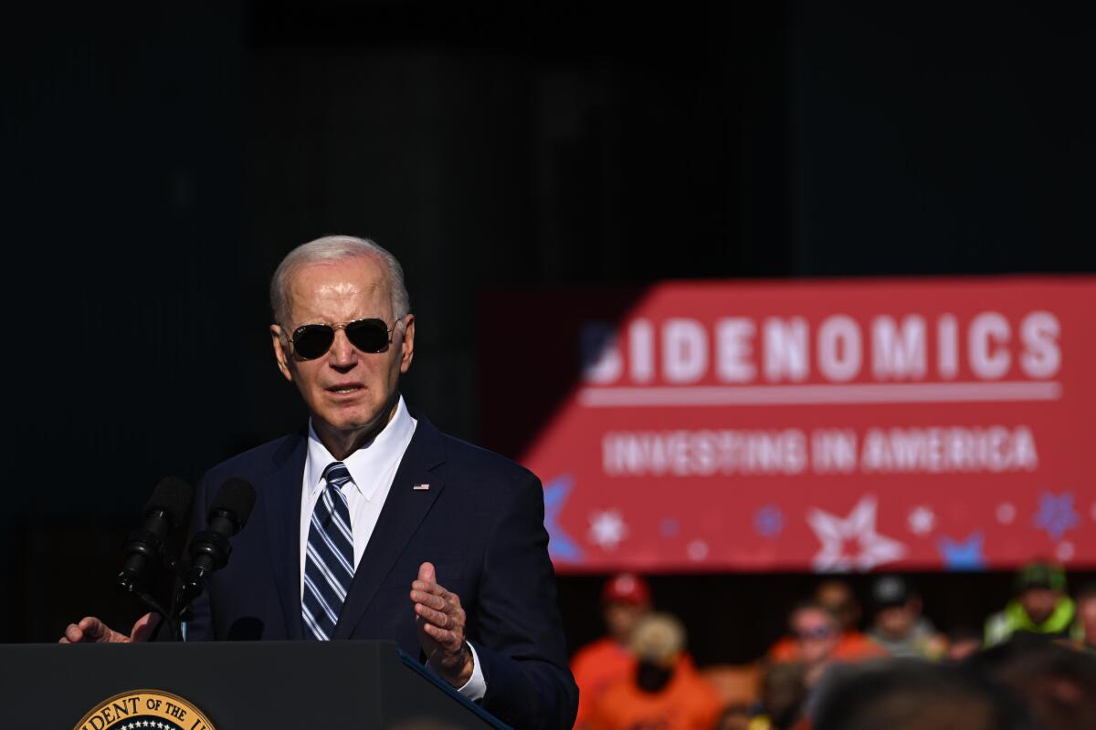 President Biden, wearing aviator sunglasses, speaks to a crowd before a sign that says "Bidenomics"