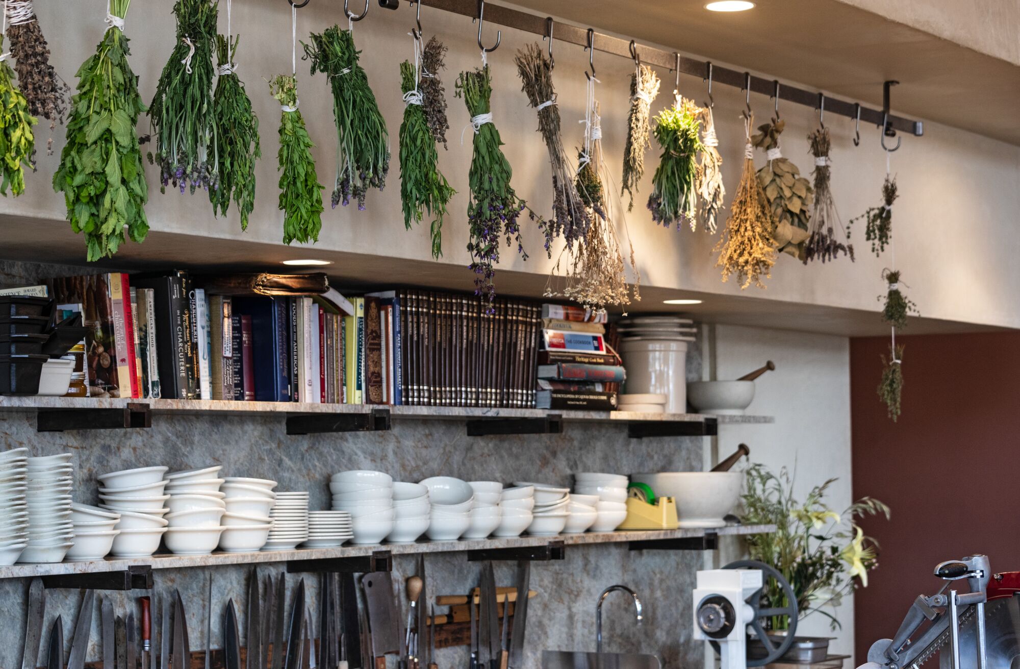 Bunches of herbs hang above shelves lined with books and dishes.