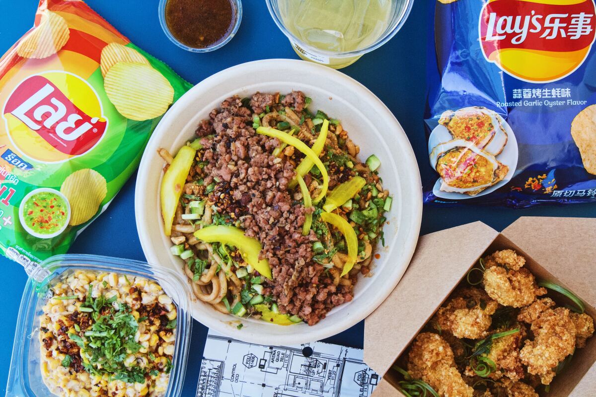 An overhead of a bowl of dan dan noodles, a box of popcorn chicken, and bags of Asian potato chips at Gingergrass Mini Mart