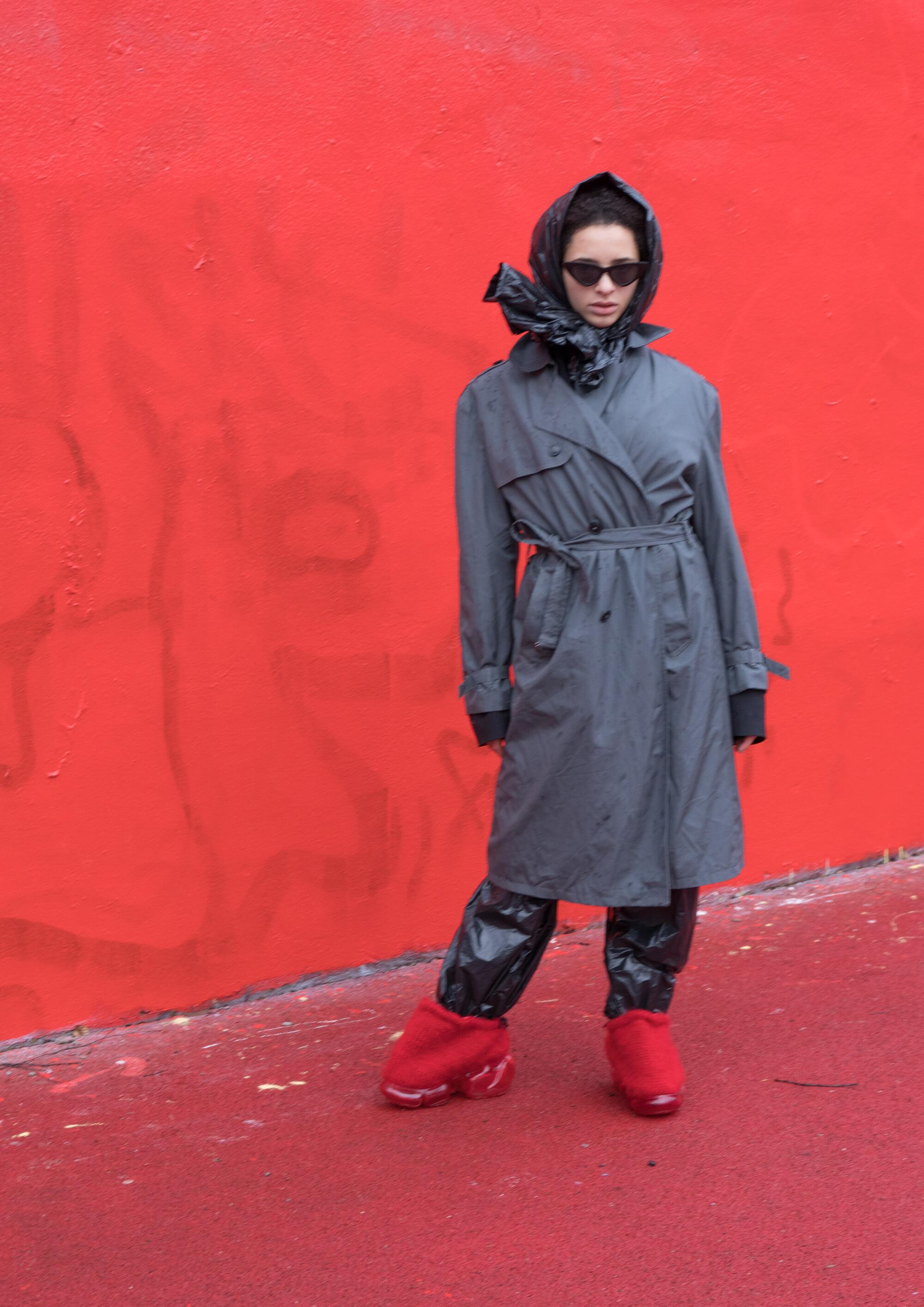 a model standing in front of a red wall wearing a gray trench coat and red grounds and H. Lorenzo shoes