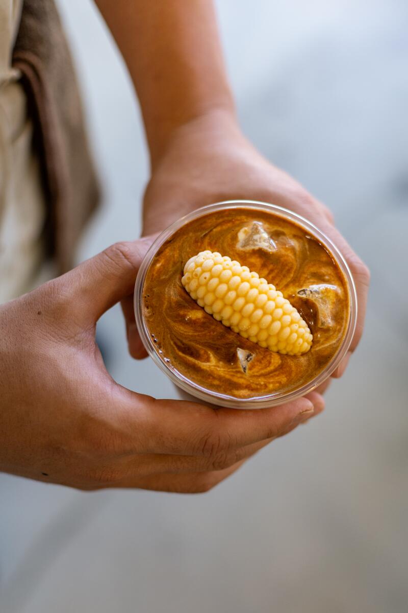 Hands hold a round container of a brown-frosted cake with a molded ear of corn on top