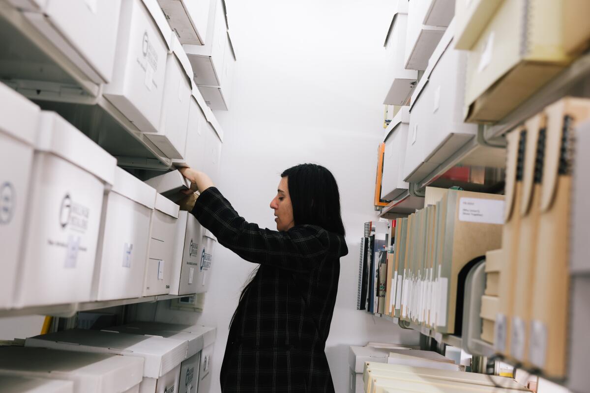 A woman in black stands between two rows of shelves filled with boxes and folders, reaching into a cardboard box.