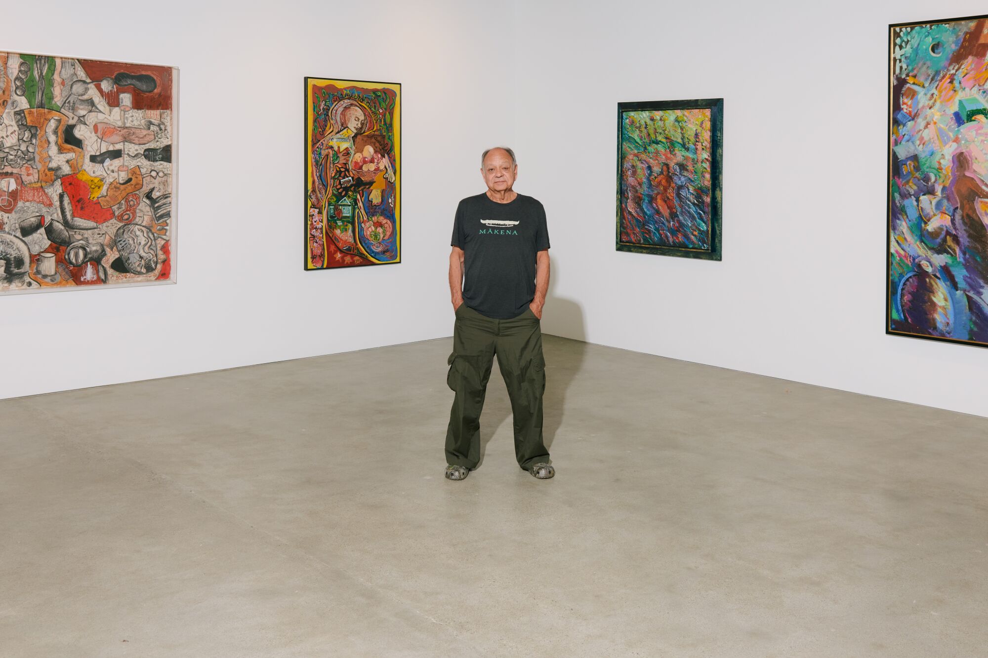 A man stands with his hands in his pockets in front of colorful paintings in a white-walled gallery.