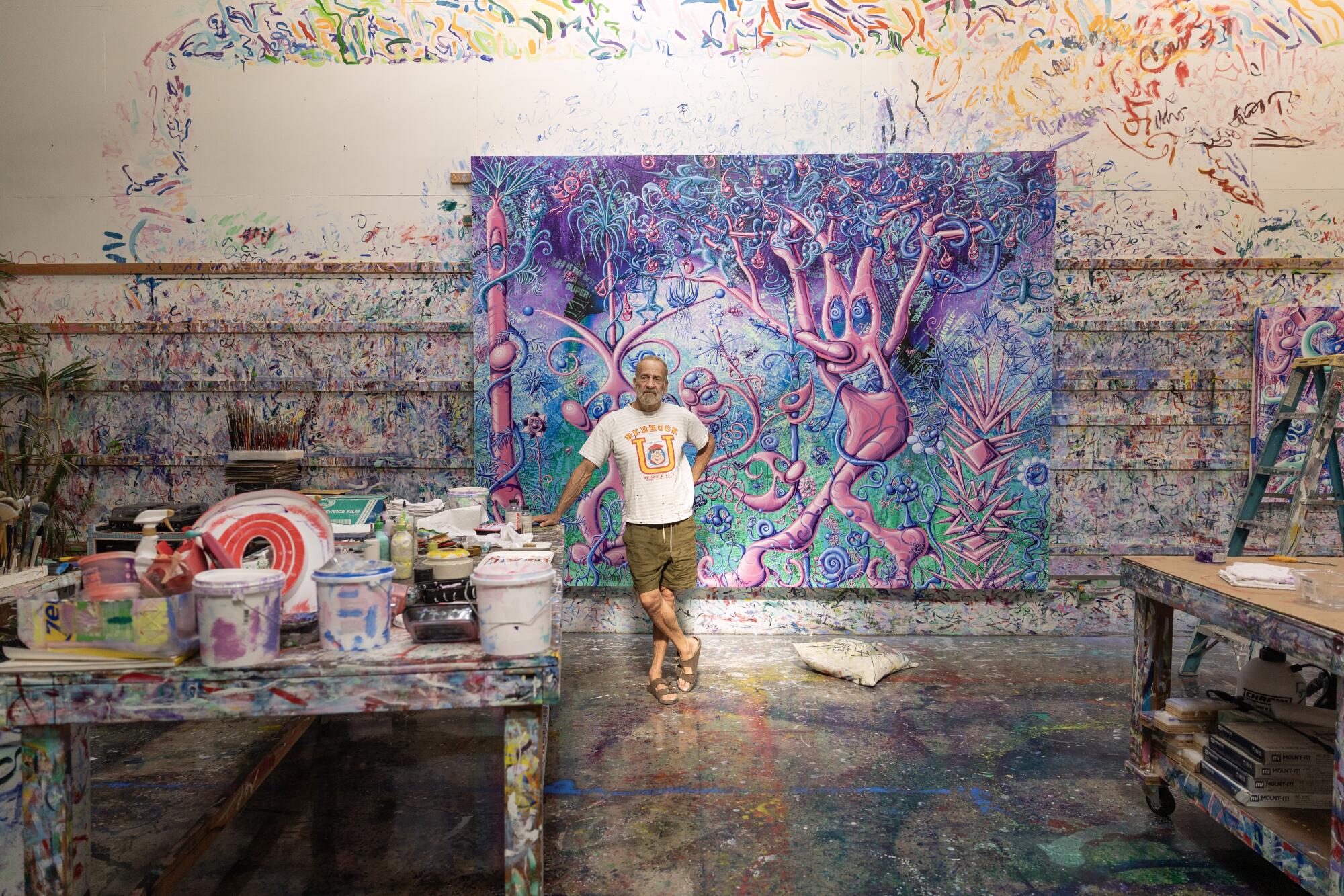 A tinta cobre as paredes, o chão, as mesas e uma grande tela atrás de Kenny Scharf, vestindo camiseta e shorts.