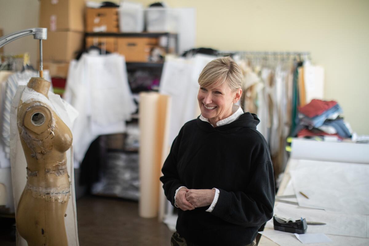 A smiling Mary Price surrounded by a garment mannequin, fabric on hangers and a table with cutting room tools