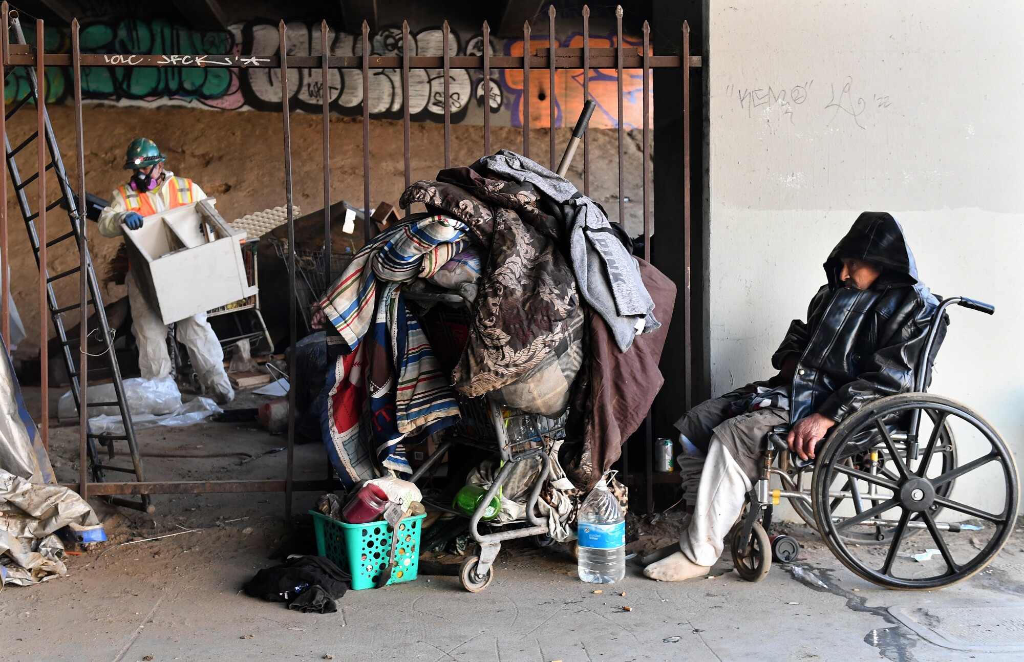 A homeless man sits in a wheelchair next to a cart of personal belongings. 