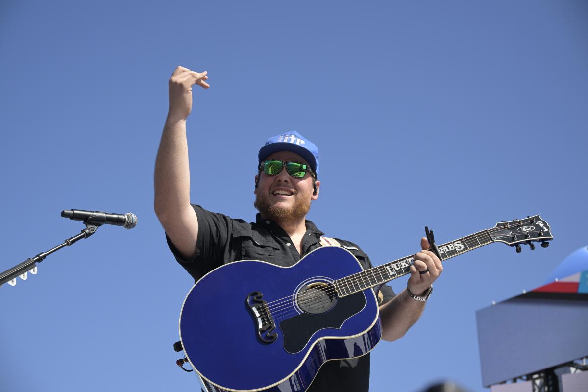 A man in a blue hat and sunglasses holding a blue acoustic guitar and raising his right hand in front of a microphone