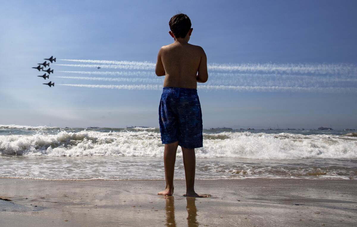 Contrails extend across the sky as a boy in swim trunks stands on a beach and watches jets passing in formation.