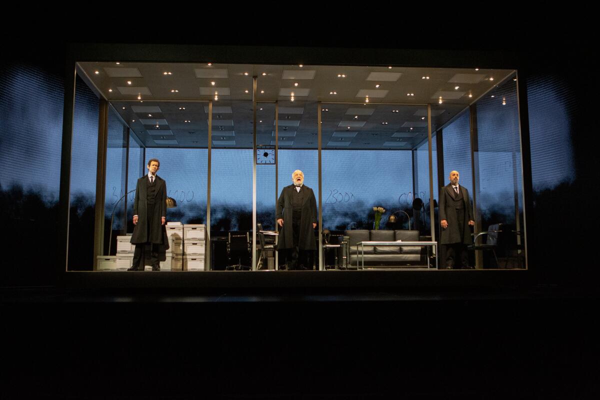 In "The Lehman Trilogy" at the Ahmanson Theatre, three men stand in a minimalist office set against a video screen cityscape.